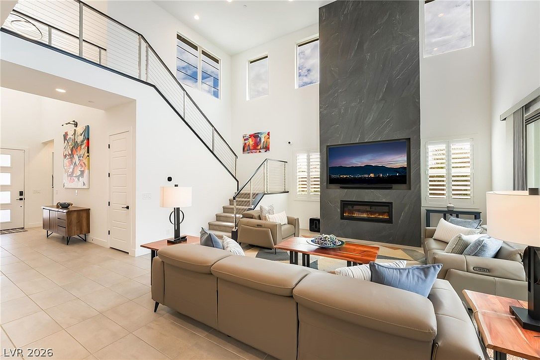 This spacious, open-concept living room features a dramatic, dark stone-clad chimney breast that serves as a focal point for the space. The room is anchored by a large, neutral-toned sectional sofa and a wooden coffee table, set against a backdrop of tall windows that allow for abundant natural light. A modern staircase with a metal railing leads to an upper landing, contributing to the airy, high-ceilinged atmosphere of this contemporary home.