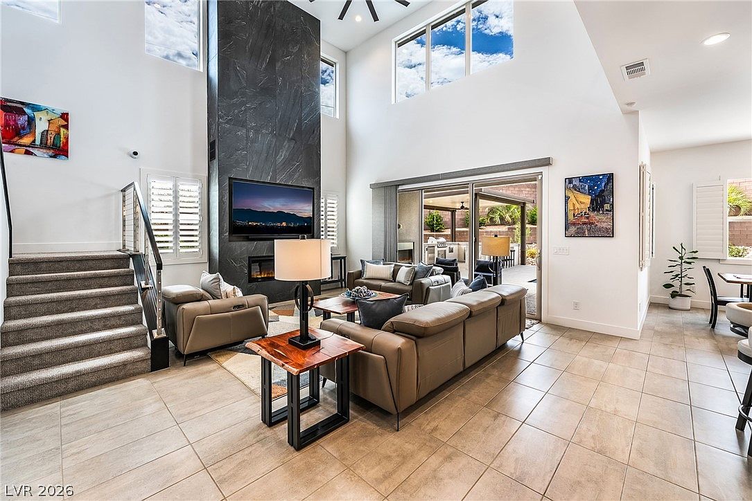This spacious, open-concept living room features a dramatic, dark stone-clad chimney breast that serves as the focal point of the room. The space is anchored by a large, neutral-toned sectional sofa and a live-edge wood side table, with a staircase leading to an upper level on the left. Large windows and a sliding glass door provide ample natural light and a seamless transition to the outdoor patio area.