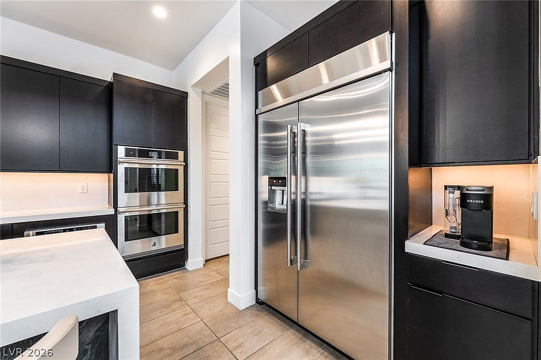 This modern kitchen features sleek black cabinetry paired with light-toned stone countertops and stainless steel appliances, including a double wall oven and a large side-by-side refrigerator. A coffee maker sits on the counter, and the space is illuminated by under-cabinet lighting, creating a sophisticated and functional atmosphere. The perspective captures the clean lines of the cabinetry and the transition into an adjacent hallway, highlighting the contemporary design.