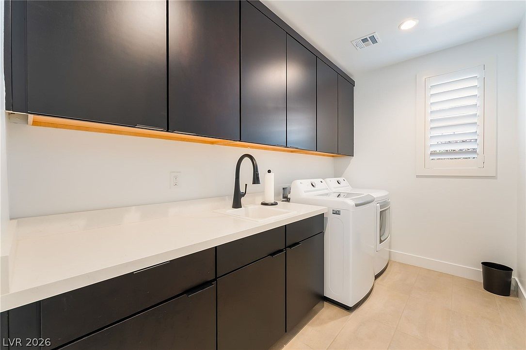 This modern laundry room features sleek, dark cabinetry paired with a light-colored countertop and an integrated sink. The space includes a side-by-side washer and dryer set, a window with plantation shutters, and under-cabinet lighting that adds a warm glow to the workspace. The room is clean, functional, and designed with a contemporary aesthetic.