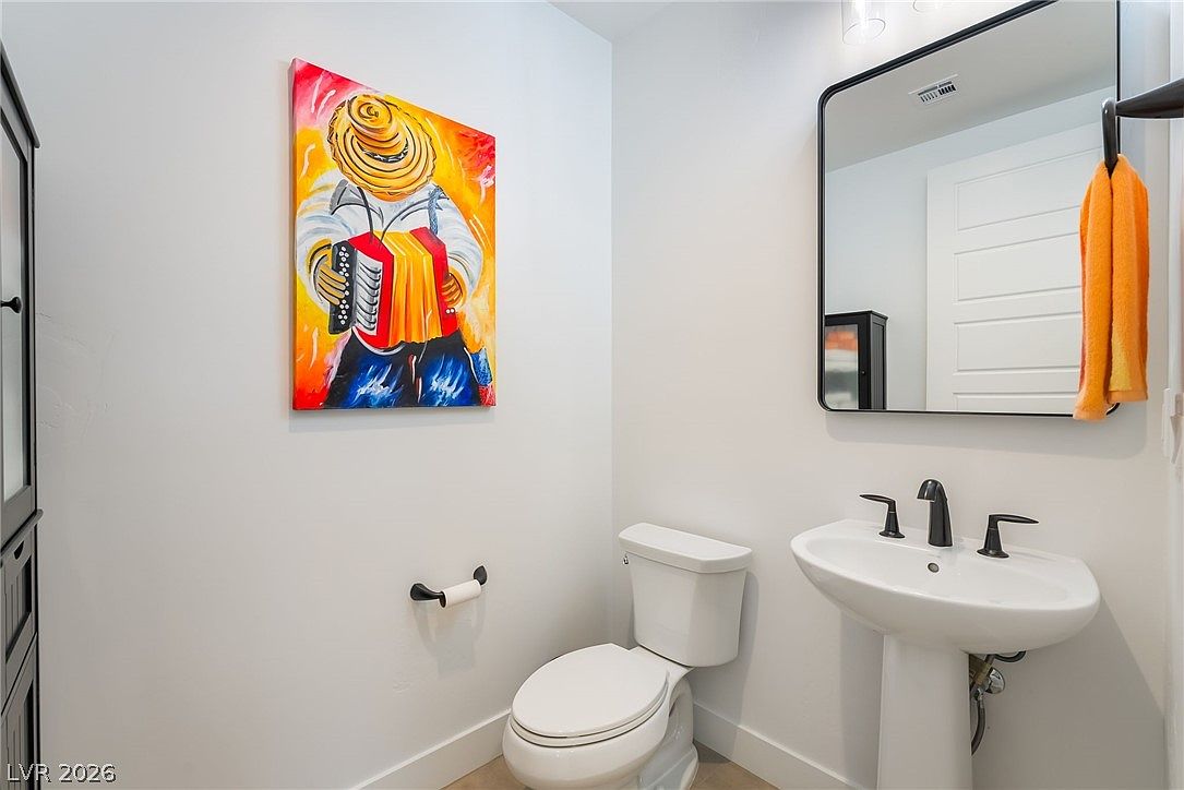 This compact powder room features a white pedestal sink with a modern dark-toned faucet and a matching rectangular mirror above. A vibrant, colorful painting of a musician adds a bold artistic focal point to the neutral-toned walls. The space is completed by a standard white toilet and a wall-mounted toilet paper holder, creating a clean and functional guest bathroom.