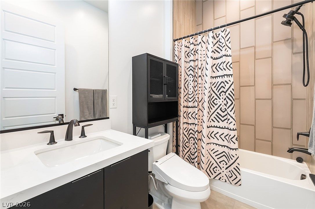 This modern bathroom features a sleek vanity with a white countertop and dark cabinetry, complemented by a large mirror and contemporary fixtures. A shower-tub combination is enclosed by a patterned shower curtain and features light-toned vertical wall tiles. A black metal storage unit sits above the toilet, adding a functional and industrial touch to the clean, minimalist space.
