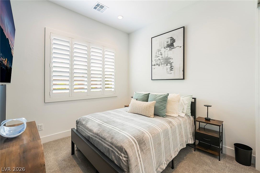 This guest bedroom features a neutral color palette with light gray walls and plush carpeting, creating a calm and inviting atmosphere. The room is furnished with a bed dressed in striped bedding, accented by sage green and white pillows, and a dark metal-framed nightstand. A large piece of abstract wall art hangs above the bed, while white plantation shutters on the window allow for adjustable natural light.