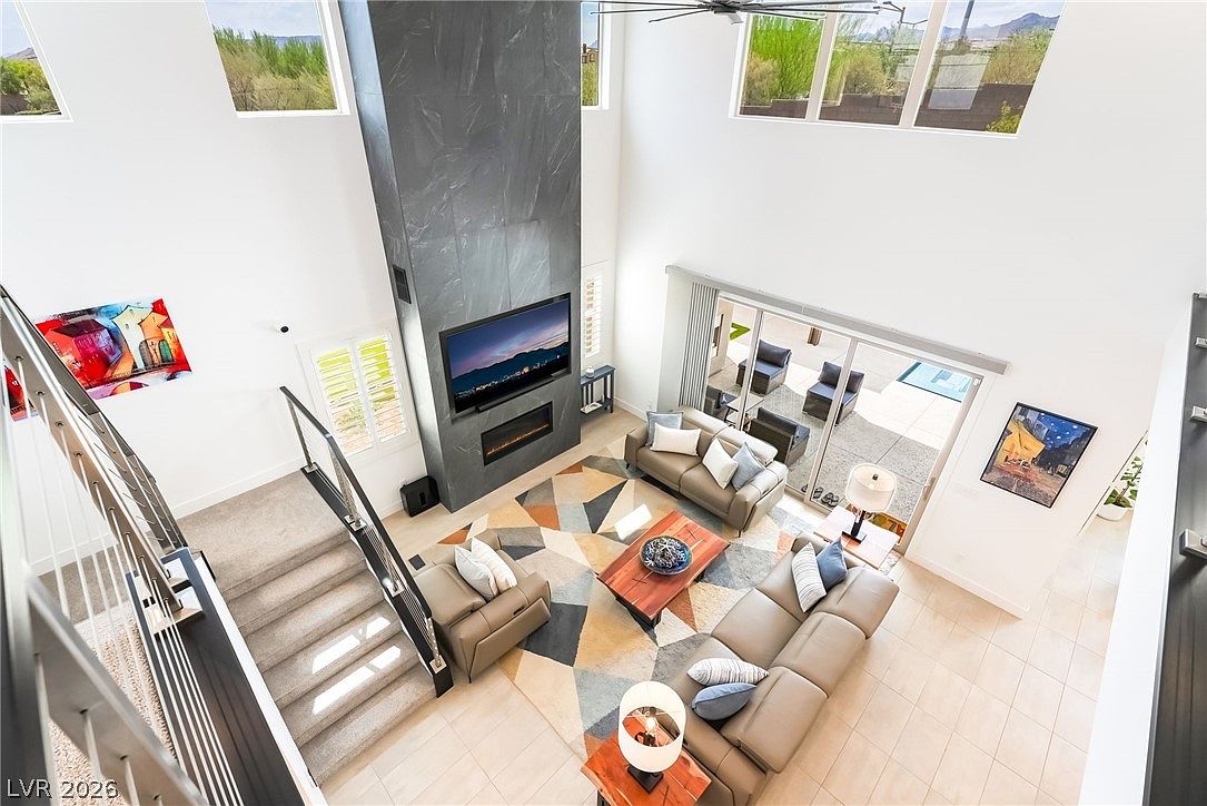 This high-angle shot captures a spacious, open-concept living room with a dramatic, tall dark stone chimney breast as the central focal point. The room features a modern seating arrangement with a leather sofa and armchairs centered around a geometric area rug and a wooden coffee table. Large sliding glass doors open to an outdoor patio, while tall windows above allow natural light to flood the airy, double-height space.