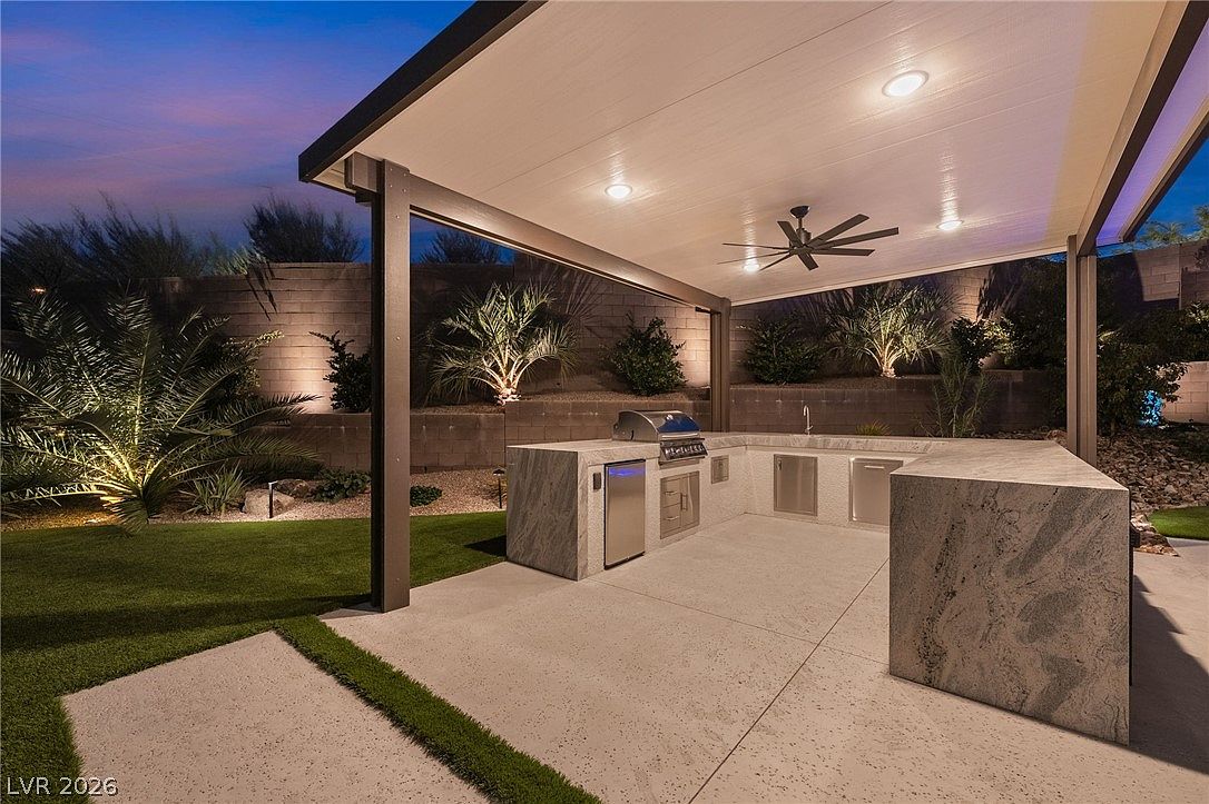 This outdoor living space features a covered patio area equipped with a built-in stone-clad kitchen island, complete with a grill, sink, and stainless steel appliances. The area is illuminated by recessed lighting and a ceiling fan, creating a comfortable environment for evening entertaining. The patio overlooks a landscaped backyard with artificial turf and decorative stone walls, offering a polished and modern aesthetic.
