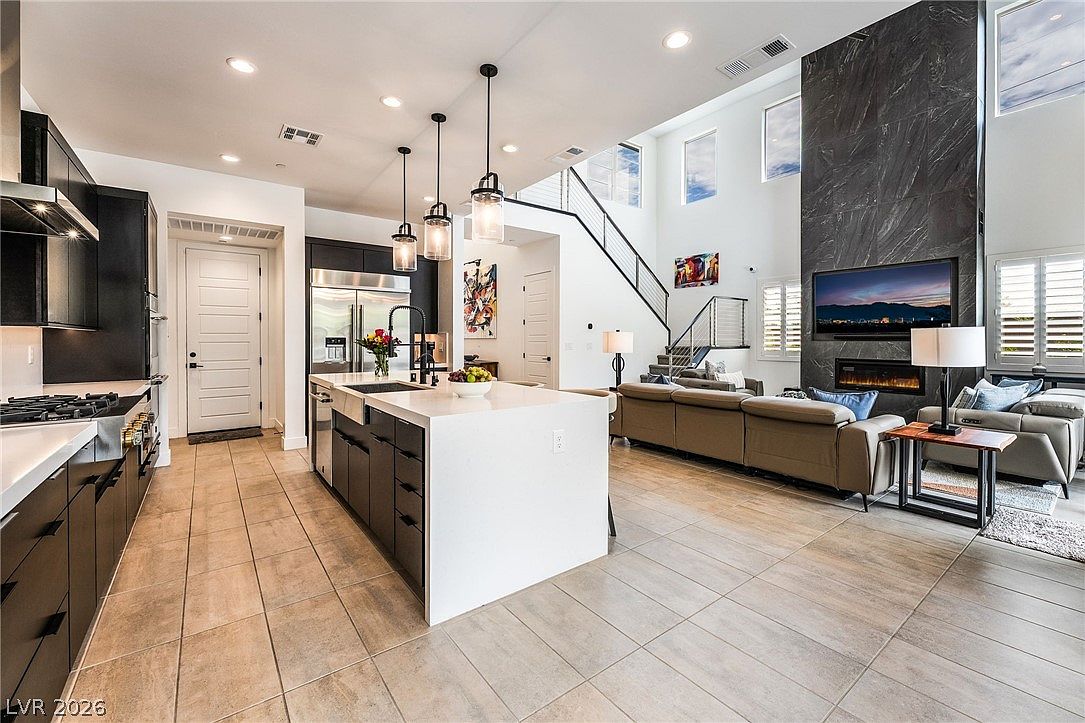 This open-concept living space features a modern kitchen with dark cabinetry, a large central island with a light-toned countertop, and three pendant lights. The kitchen flows seamlessly into a spacious living area anchored by a dark stone-clad chimney breast and a large television. The room is characterized by high ceilings, neutral tile flooring, and a staircase with a metal railing, creating an airy and contemporary atmosphere.