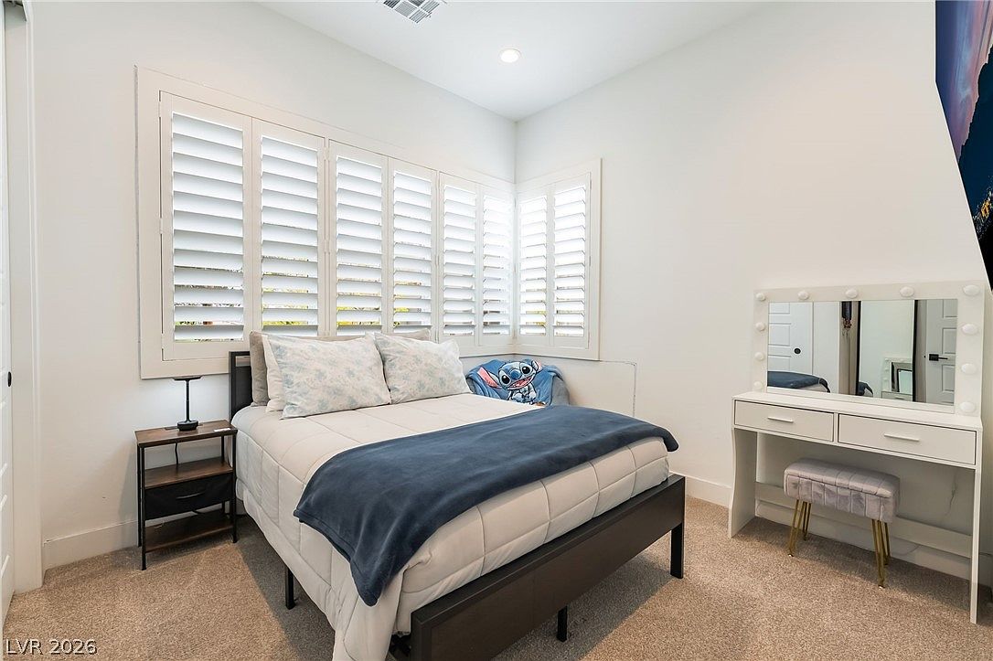 This cozy bedroom features a dark-framed bed dressed in white bedding with a navy blue throw blanket. To the right, a white vanity with a lighted mirror and a matching upholstered stool provides a dedicated space for grooming. The room is illuminated by recessed lighting and natural light filtering through white plantation shutters covering the corner windows.