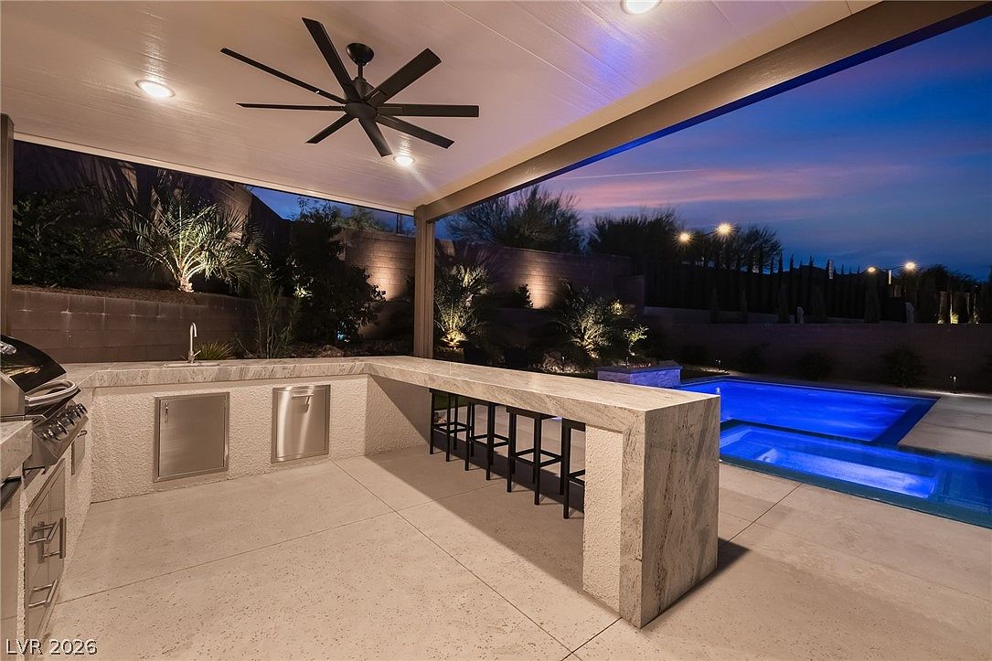 This outdoor living space features a covered patio equipped with a built-in kitchen, including a grill, sink, and a stone-topped bar with seating. The area is illuminated by recessed lighting and a large ceiling fan, creating a comfortable transition to the adjacent swimming pool and spa. The scene is captured at twilight, highlighting the blue glow of the water and the landscaped backyard.