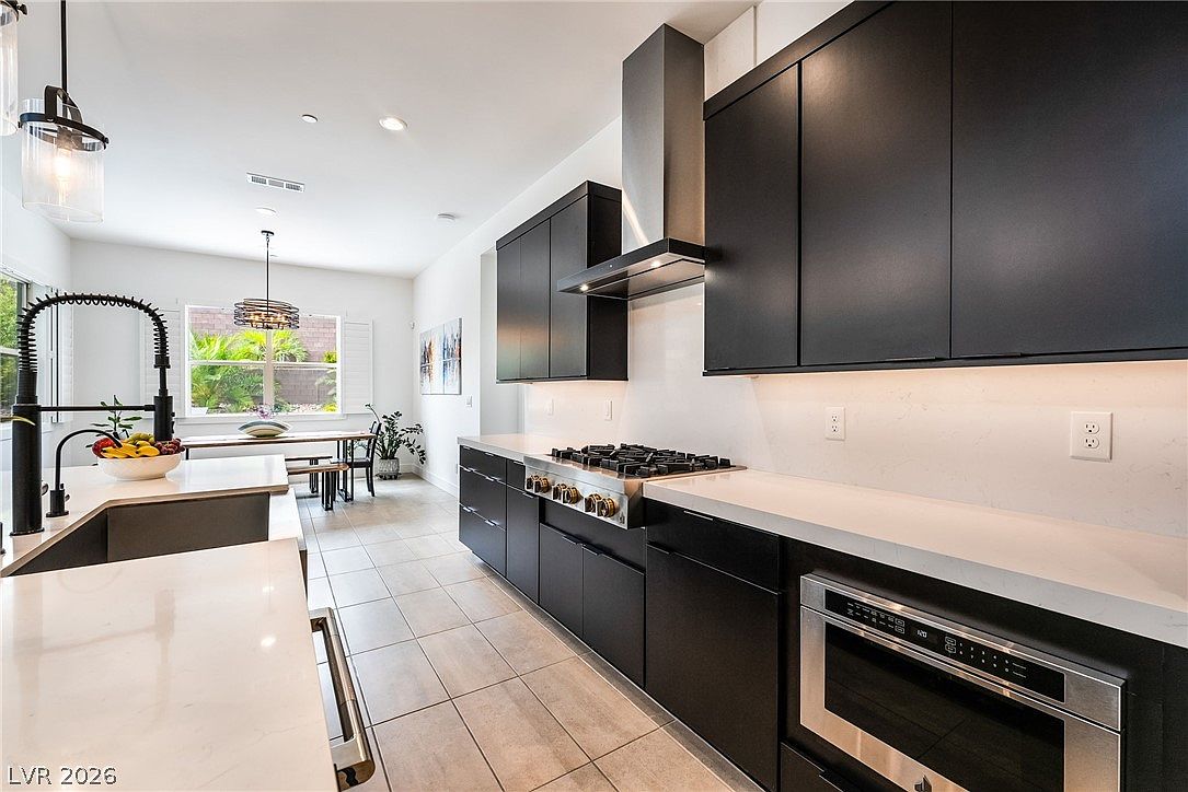 This modern kitchen features sleek matte black cabinetry contrasted by light-toned stone countertops and a matching backsplash. A stainless steel gas range with a professional-style vent hood serves as the focal point, while a large island with a farmhouse-style sink provides additional workspace. The open-concept layout flows seamlessly into a bright dining area, creating a cohesive and contemporary living space.