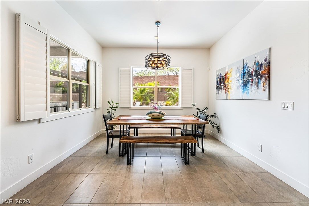 This bright and airy dining room features a rustic wood-slab table paired with matching benches and two black chairs, centered under a modern, spherical metal pendant light. The space is framed by large windows with white plantation shutters that allow natural light to flood the room, highlighting the neutral tile flooring. Three abstract, colorful canvas prints hang on the right wall, adding a vibrant focal point to the clean, minimalist aesthetic.