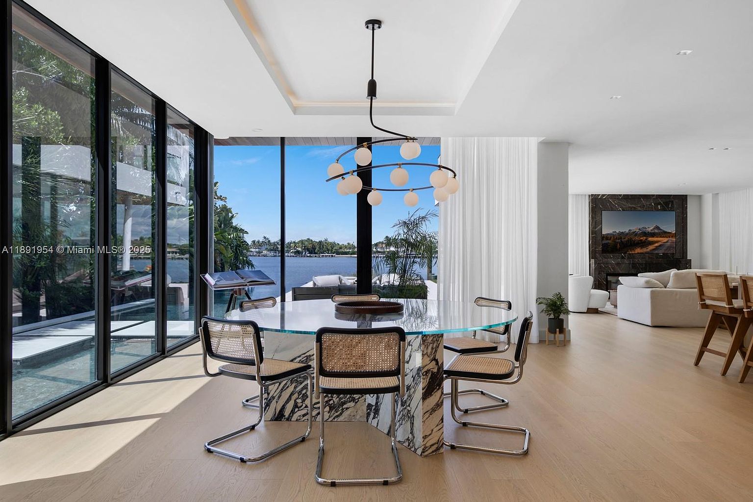 This interior shot showcases a modern dining room with a round glass-top table and unique chairs with chrome frames and woven backs. The room features large windows offering a view of the water, a stylish chandelier, and light wood flooring. The space is bright and airy, creating an inviting atmosphere for dining and entertaining.