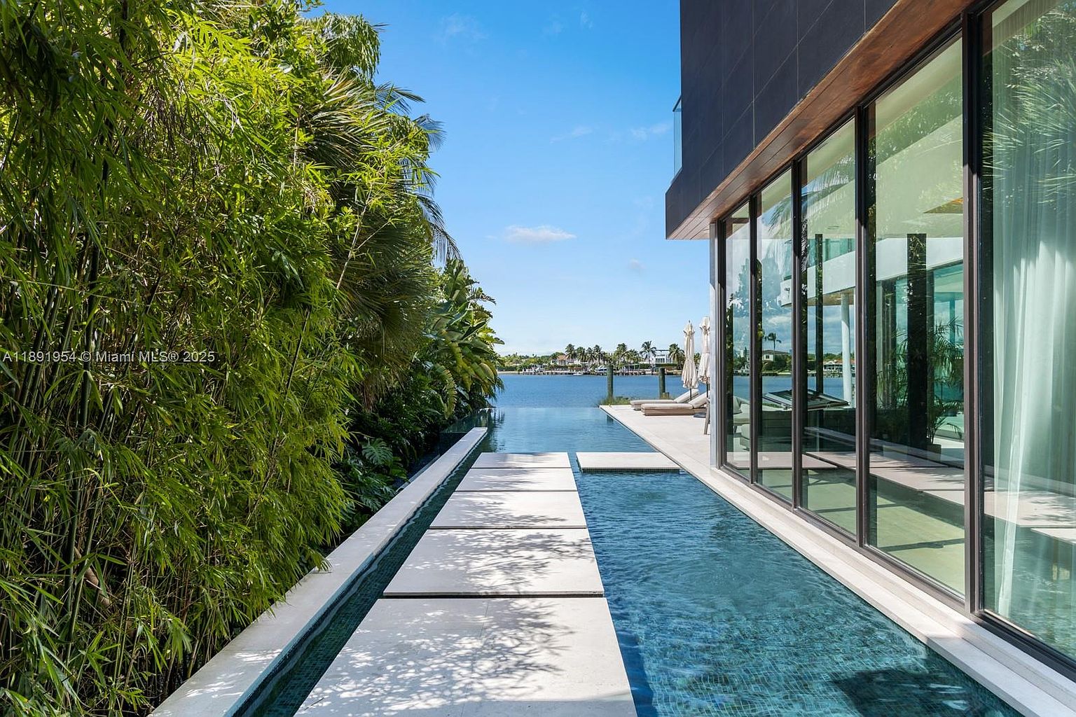 This image showcases a luxurious outdoor pool area, featuring a modern architectural design with floor-to-ceiling windows and a sleek, dark exterior. A narrow pool runs alongside the building, with stepping stones leading towards a waterfront view. Lush greenery provides privacy and enhances the serene atmosphere, creating an inviting and upscale outdoor living space.