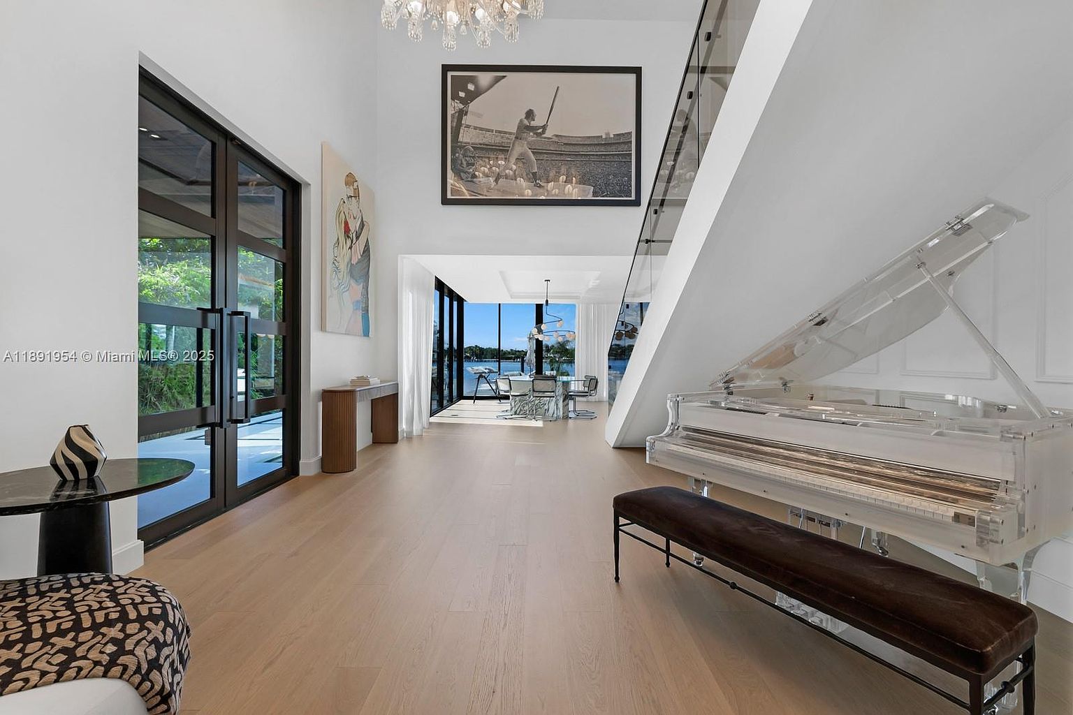 This interior shot showcases a grand foyer with high ceilings and an open floor plan. A clear grand piano sits elegantly near a modern staircase with glass railings, while a large black and white photograph hangs above the entryway to the dining area. The space is bright and airy, emphasizing luxury and contemporary design.