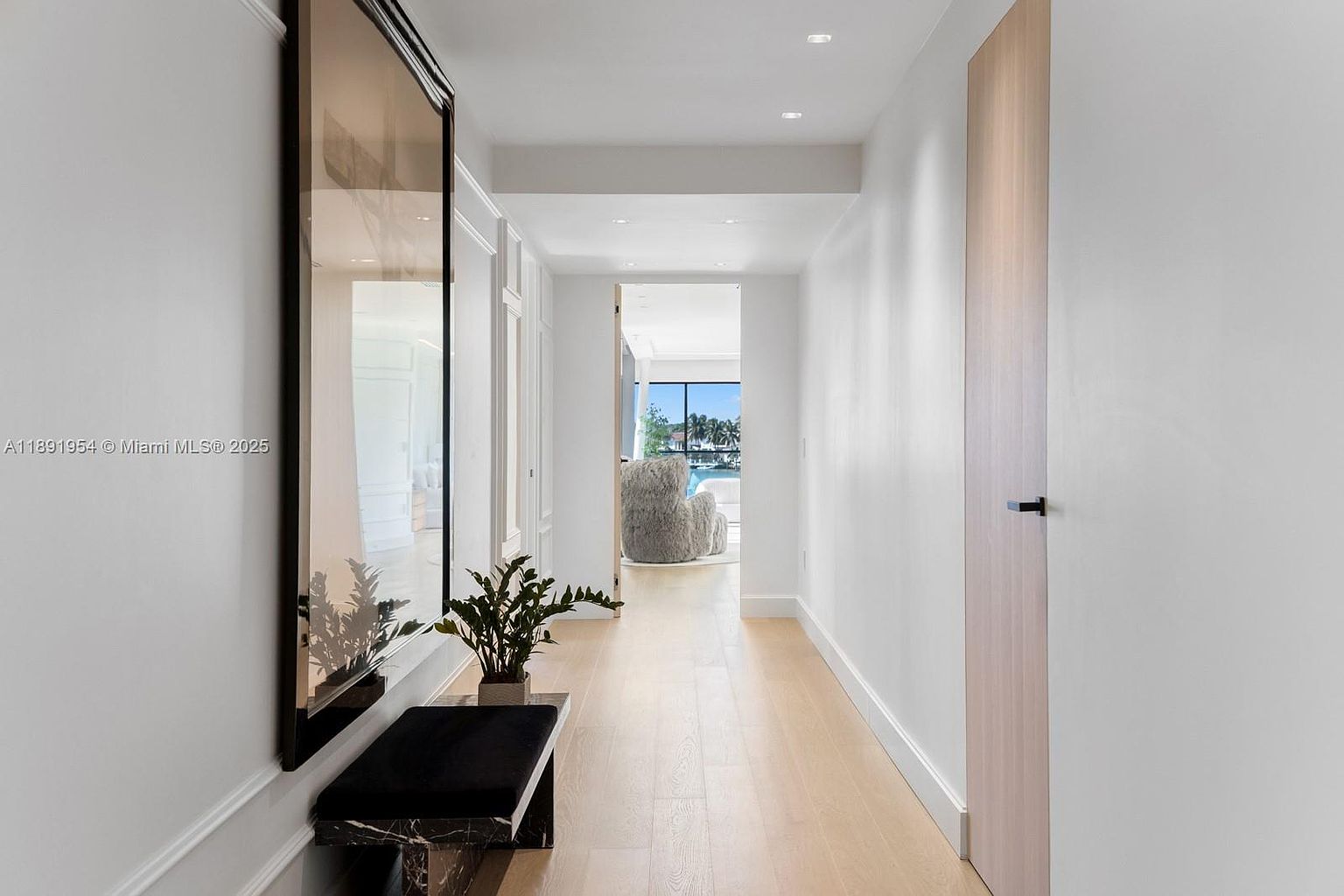 This interior shot showcases a bright and modern hallway with light wood flooring and white walls. A large framed mirror and a sleek bench with a plant add a touch of elegance, while a doorway at the end of the hall offers a glimpse into another room with a view of the water. The overall impression is clean, spacious, and sophisticated.