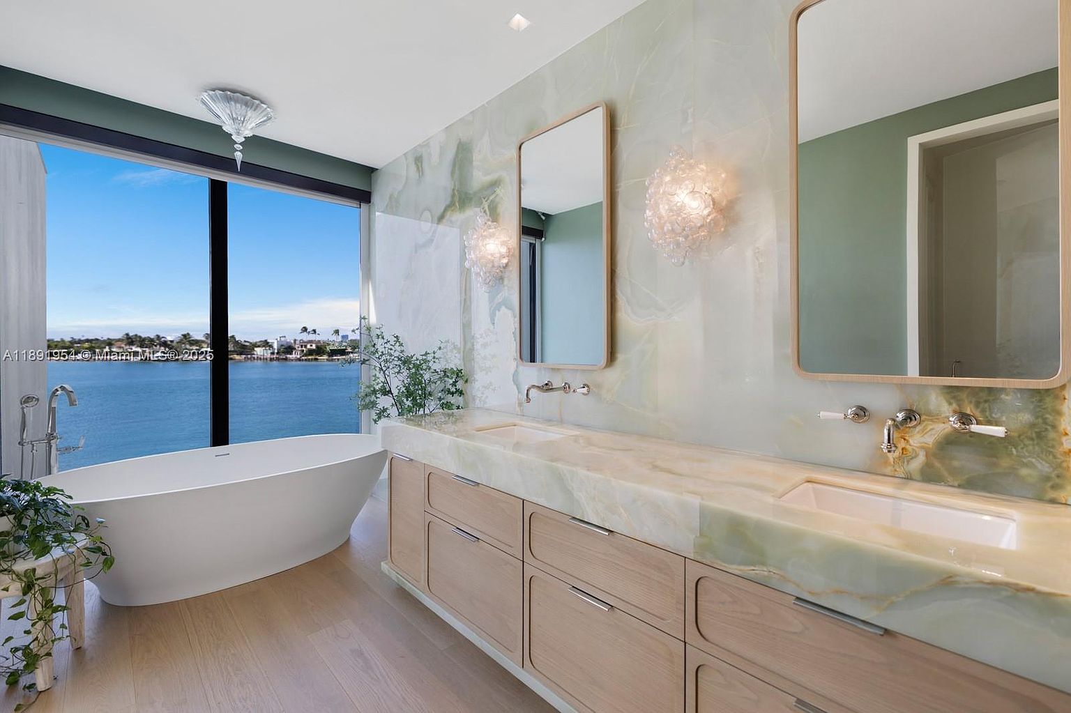 This is a luxurious primary bathroom featuring a freestanding soaking tub positioned by a large window with a waterfront view. The vanity area showcases a light wood cabinet with a marble countertop and dual sinks, complemented by elegant mirrors and sconces. The overall design exudes a spa-like ambiance with its natural light and high-end finishes.