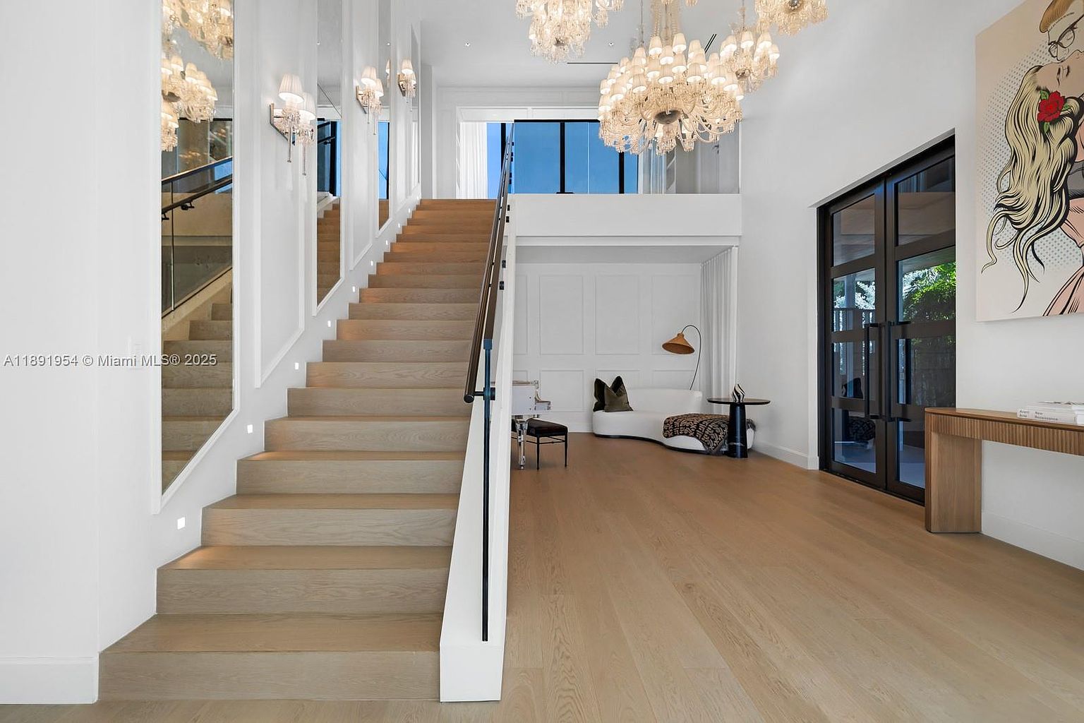 This interior shot showcases a grand hallway with a prominent staircase. The stairs are made of light wood and feature a glass railing, complemented by white walls and modern lighting. The space is bright and airy, with a glimpse of a seating area and artwork, creating an inviting and luxurious atmosphere.