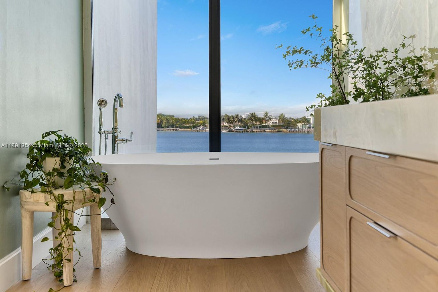 This is a luxurious primary bathroom featuring a freestanding white bathtub positioned in front of a large window with a scenic water view. The bathroom includes a modern vanity with light wood cabinetry and a plant on top, adding a touch of nature. The overall design is elegant and serene, creating a spa-like atmosphere.