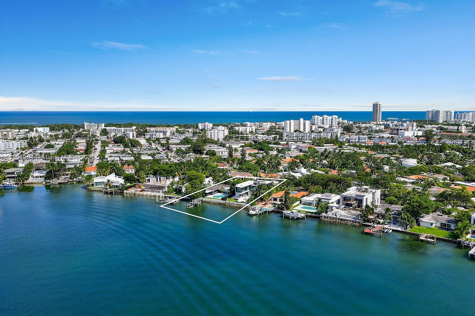 This aerial shot showcases a luxurious waterfront property with a modern architectural design. The house features clean lines, large windows, and a private pool, all situated on a canal with boat docks. The surrounding landscape includes lush greenery and a clear view of the city skyline in the distance, emphasizing the property's prime location and upscale appeal.