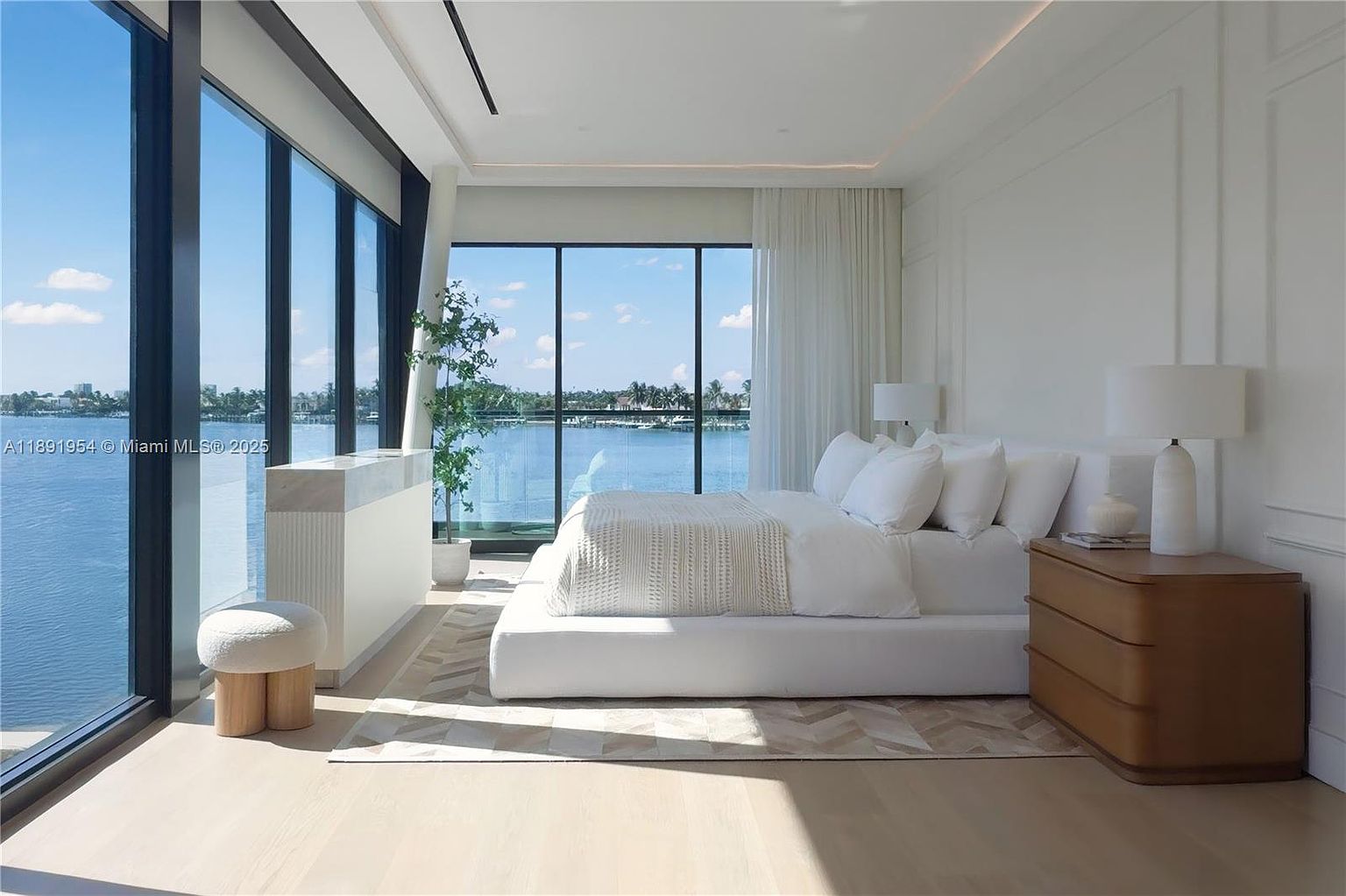 This is a bright and airy primary bedroom with floor-to-ceiling windows offering a stunning waterfront view. The room features a minimalist design with white walls, a platform bed with white linens, and a light wood floor. A modern dresser and a stylish stool add to the room's sophisticated and serene atmosphere.