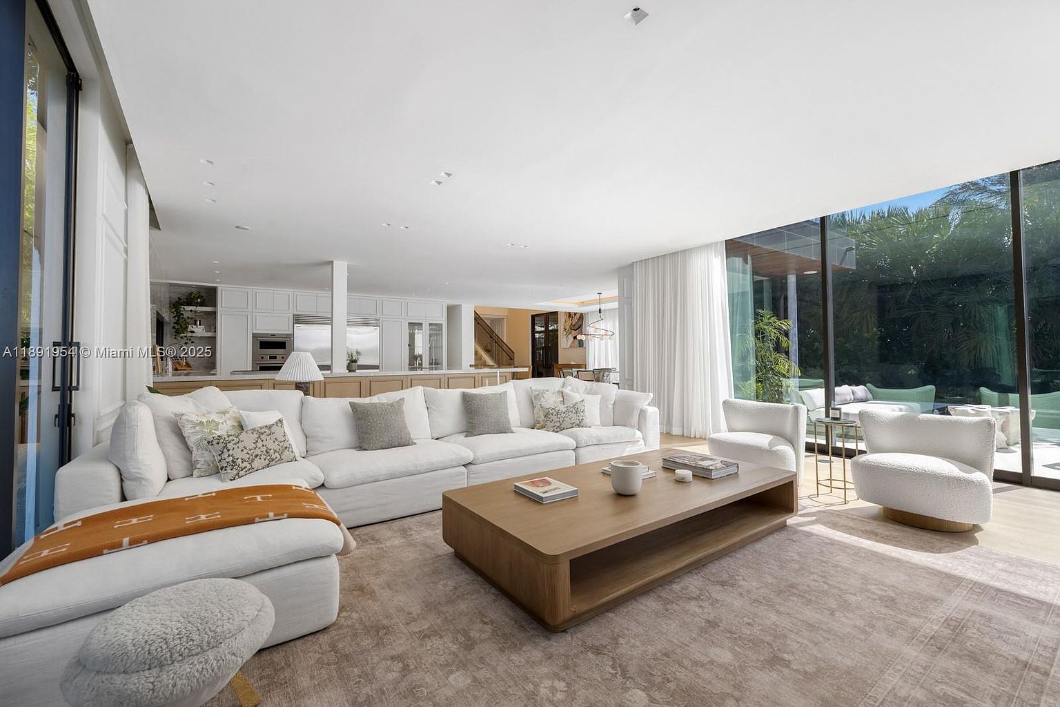This is a bright and spacious living room featuring a large white sectional sofa adorned with decorative pillows and a cozy throw. A substantial wooden coffee table anchors the space, complemented by two plush white armchairs. Large windows offer ample natural light and views of the outdoors, enhancing the room's airy and inviting atmosphere.