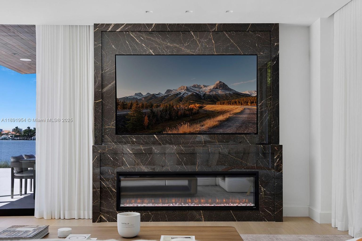 This interior shot showcases a modern living room with a striking marble-framed television and fireplace feature wall. The room is bright, with natural light filtering through sheer white curtains, highlighting the contemporary design and clean lines. A glimpse of the waterfront view adds to the luxurious ambiance.