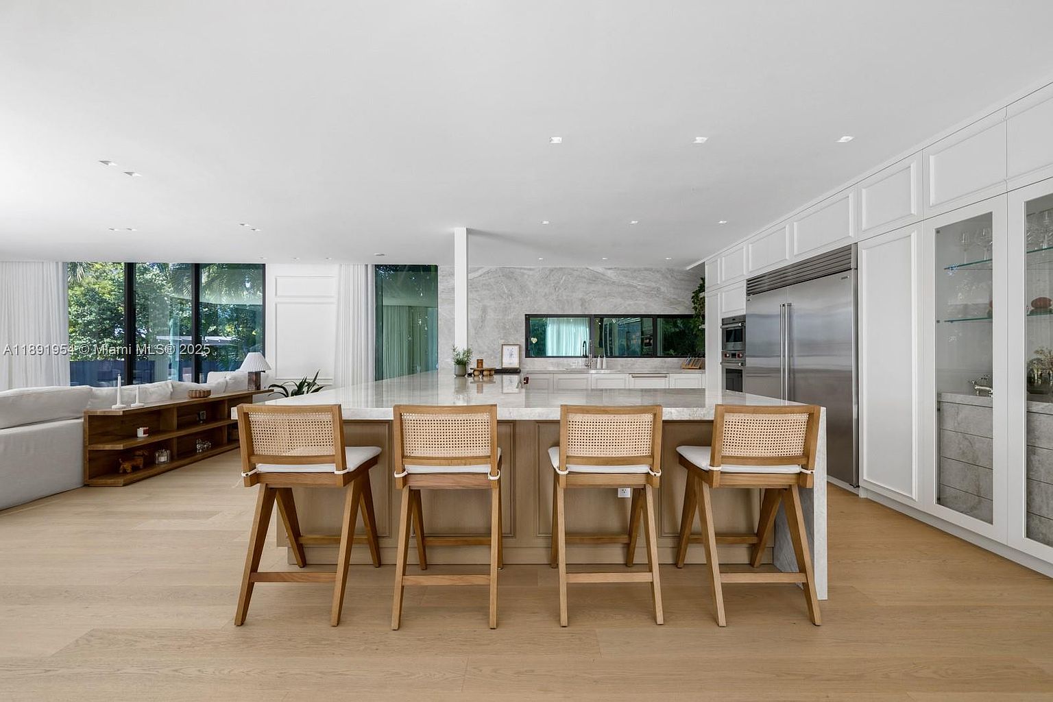 This is a bright and spacious kitchen featuring a large island with seating for four, light wood flooring, and a modern design. The kitchen includes stainless steel appliances and white cabinetry, with a large window providing natural light. The overall impression is one of luxury and functionality, perfect for a modern home.