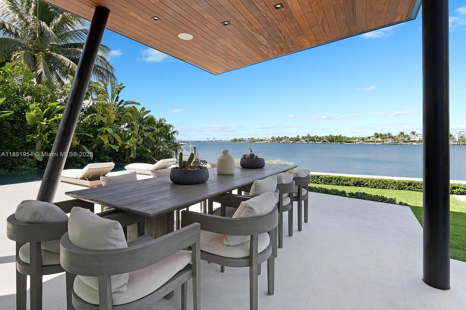 This image showcases a luxurious outdoor dining area with a stunning waterfront view. The space features a large wooden table surrounded by modern gray chairs with white cushions, all under a wooden ceiling. The scene is framed by lush greenery and a clear blue sky, creating an inviting and serene atmosphere perfect for entertaining or relaxing.