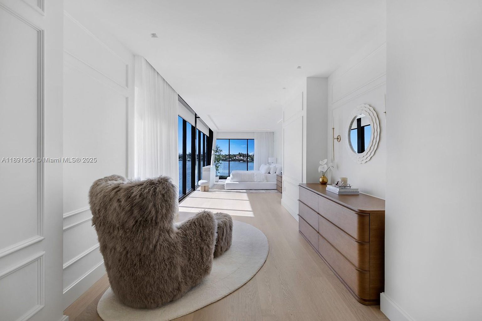 This is a bright and airy primary bedroom featuring a modern design with clean lines and a neutral color palette. The room boasts large windows offering waterfront views, a cozy fur chair with ottoman, and a sleek dresser with a round mirror above it. The overall impression is one of luxury and tranquility.