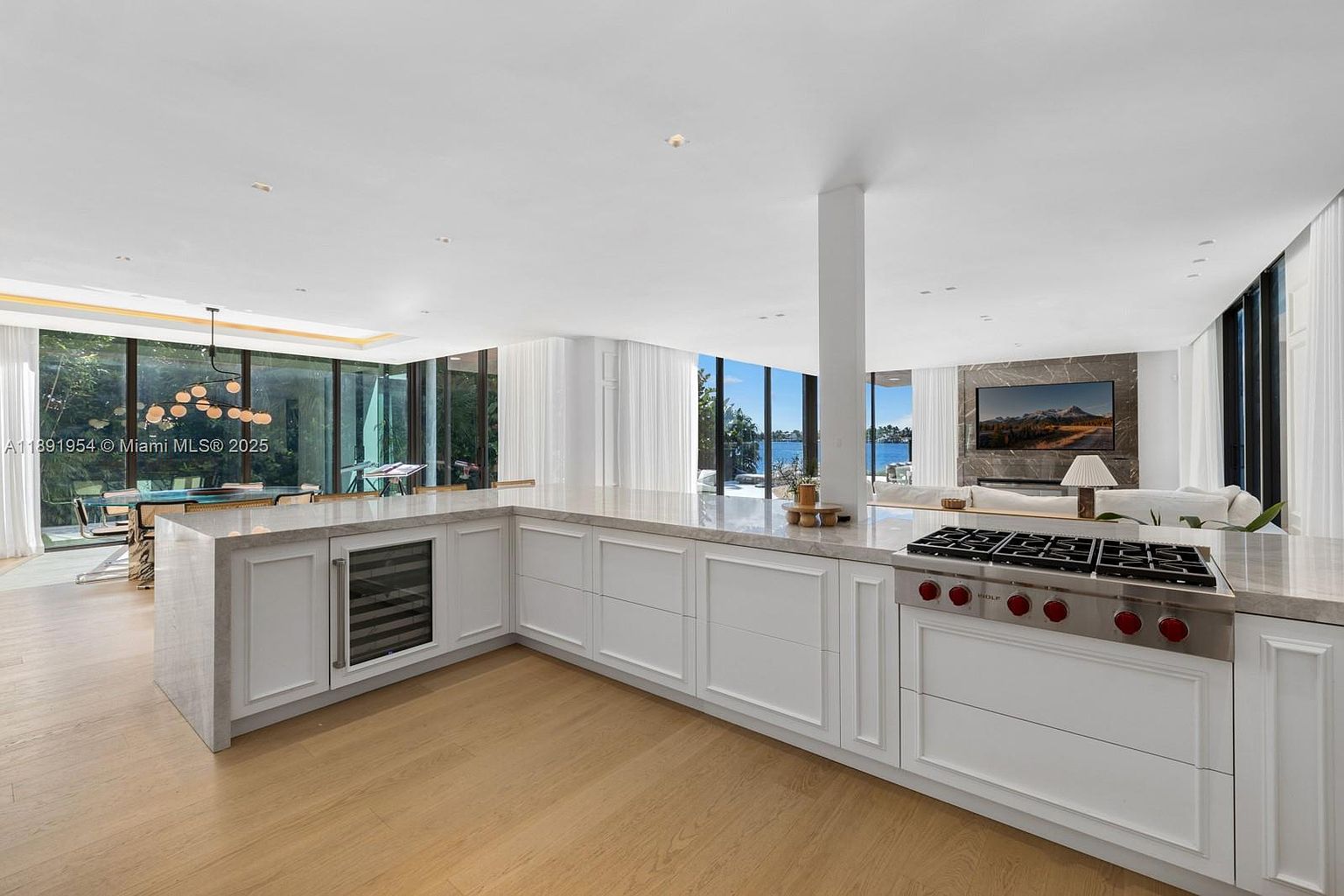 This is a bright, open-concept kitchen featuring white cabinetry, a large island with a marble countertop, and high-end stainless steel appliances. The kitchen seamlessly transitions into a living area with a fireplace and large windows offering water views. The overall impression is luxurious and modern.