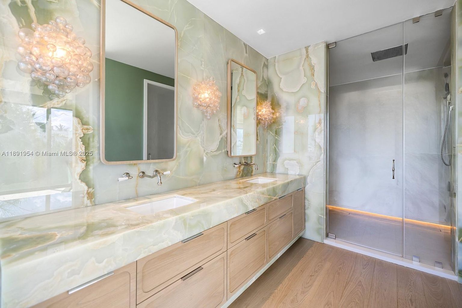 This is a luxurious primary bathroom featuring a double vanity with a light wood cabinet and a green marble countertop and backsplash. Two large mirrors and decorative sconces adorn the wall above the vanity. A glass-enclosed shower is located to the right, and the flooring is light wood. The overall impression is modern and upscale.