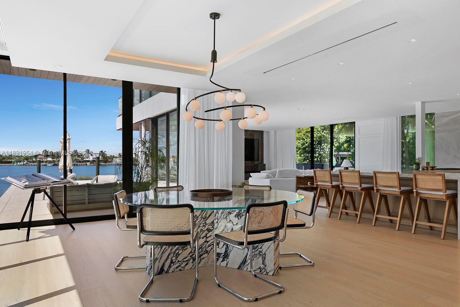 This is an interior shot of a modern dining room. The room features a round glass-top dining table with a marble base, surrounded by six chairs with chrome frames and woven seats. A contemporary chandelier hangs above the table, and large windows offer a view of the waterfront, creating a bright and airy atmosphere.