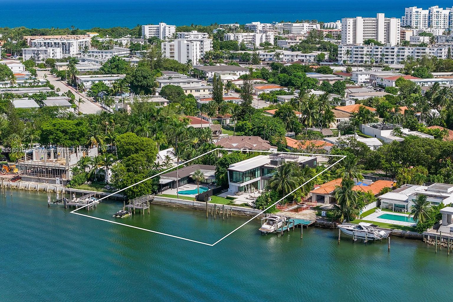 This aerial shot showcases a modern waterfront property with clean architectural lines and expansive glass walls. The property features a pool, a private dock with boats, and lush landscaping. The view extends to the ocean and the city skyline, emphasizing the property's prime location and luxurious appeal.