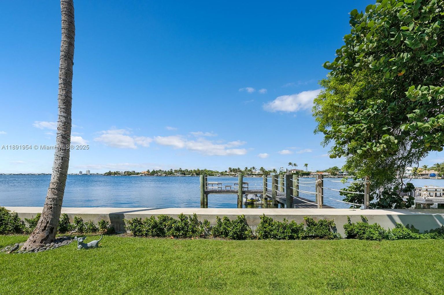 This exterior shot showcases a well-maintained yard with lush green grass and manicured hedges, leading to a waterfront view. A wooden dock extends into the calm water, creating a serene and inviting atmosphere. The presence of a mature tree and a palm tree adds to the tropical feel of the property.