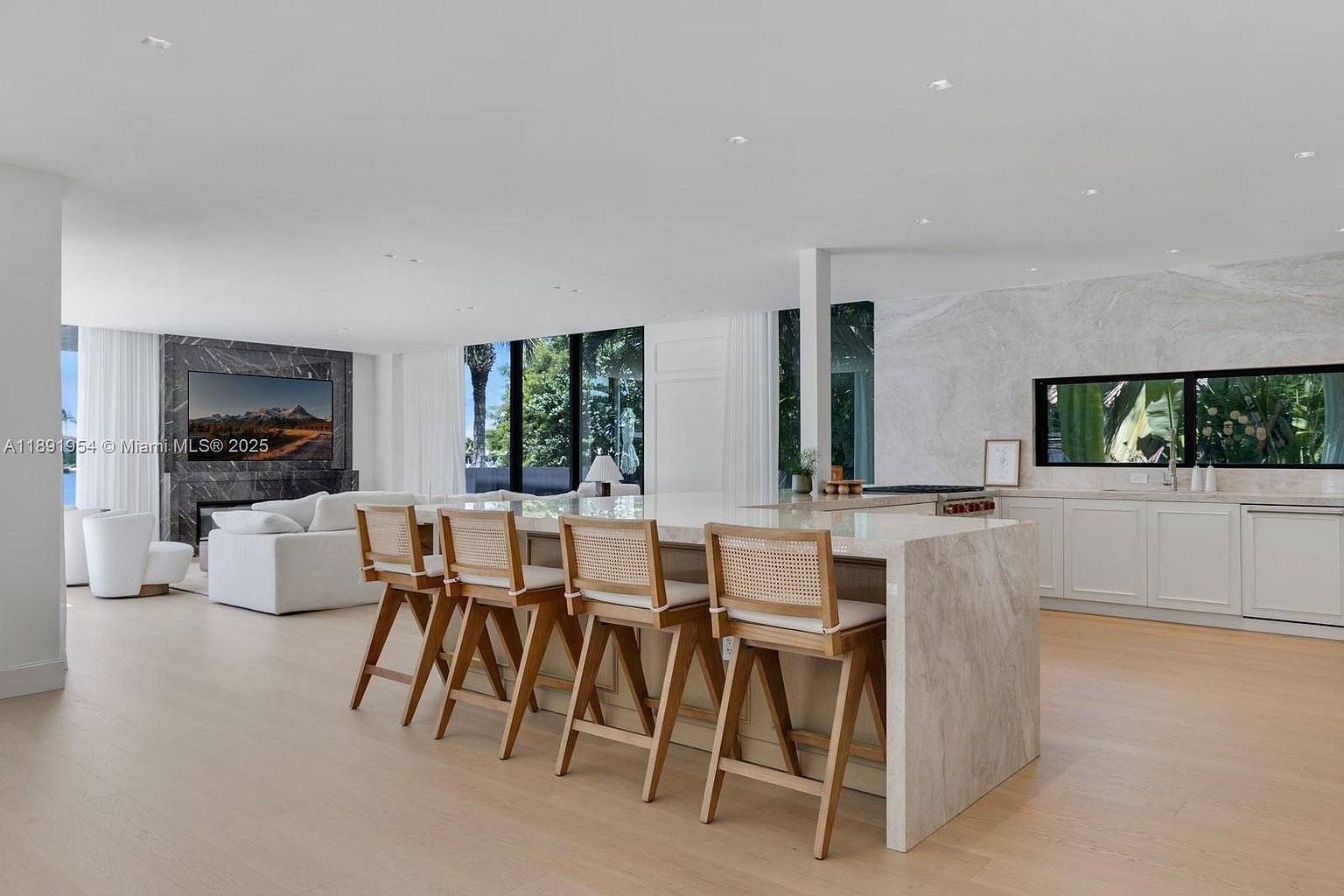 This is a bright, modern kitchen with an open floor plan that flows into the living area. The kitchen features a large island with seating, light wood floors, and white cabinetry. A marble backsplash and countertops add a touch of luxury, while large windows provide ample natural light and views of the outdoors.