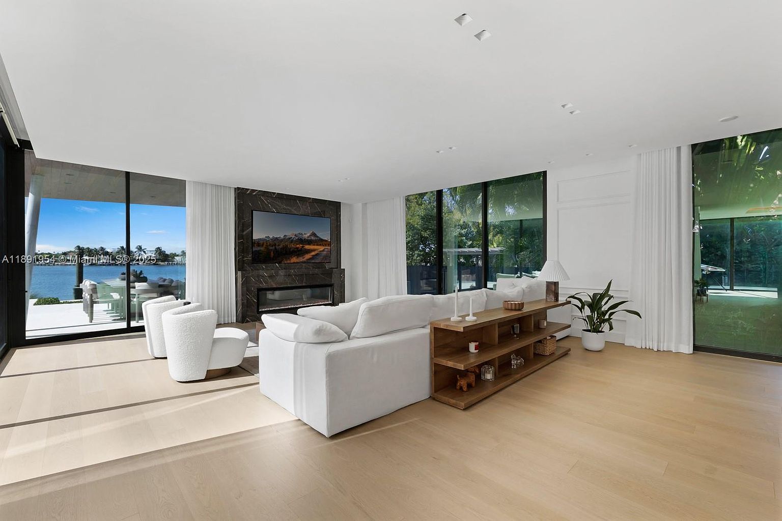 This is a bright and airy living room featuring a large white sectional sofa, modern armchairs, and a wooden shelving unit. A sleek fireplace with a dark marble surround is the focal point, complemented by large windows offering views of the outdoors. The room has a minimalist and luxurious feel, enhanced by the light wood flooring and neutral color palette.