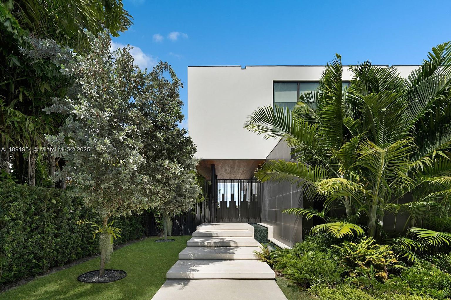 This image showcases the entryway of a modern home, featuring a minimalist design with clean lines and a neutral color palette. A series of concrete steps lead to a gated entrance, flanked by lush greenery and palm trees, creating a serene and inviting atmosphere. The architectural style emphasizes simplicity and sophistication, making a strong first impression.