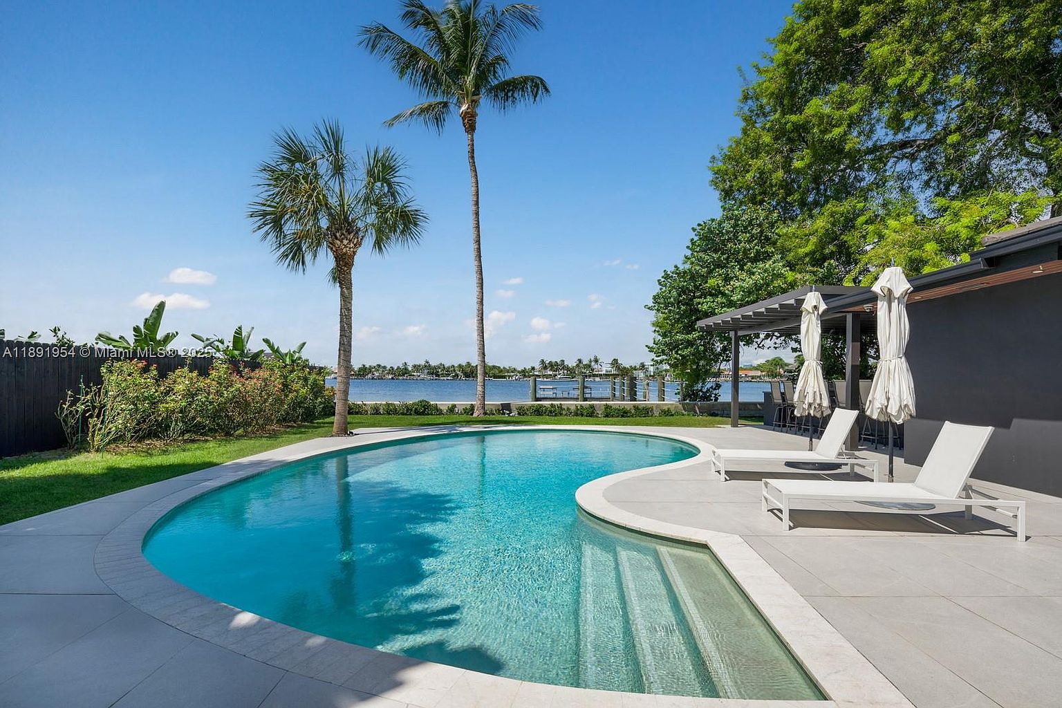 This image showcases a luxurious backyard with a freeform swimming pool as the focal point. The pool is surrounded by a light-colored patio with lounge chairs and umbrellas, offering a relaxing space. Palm trees and lush greenery add to the tropical ambiance, while the waterfront view in the background enhances the property's appeal.