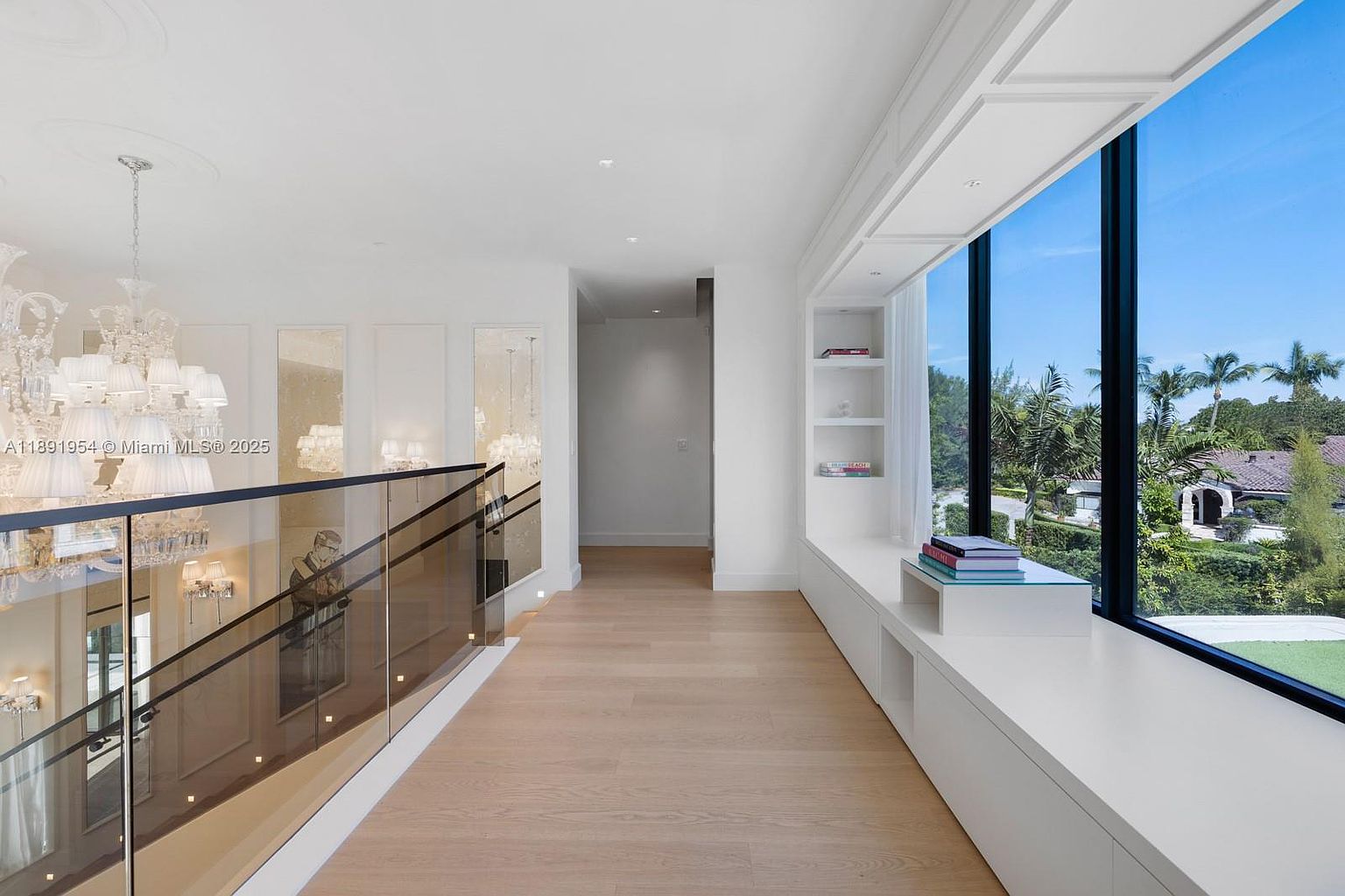 This interior shot showcases a bright and airy hallway with light wood flooring and white walls. A glass-paneled staircase with a black railing is visible on the left, while a large window with a built-in bench and shelving unit offers a view of lush greenery outside. The space is illuminated by natural light and a decorative chandelier, creating a luxurious and inviting atmosphere.