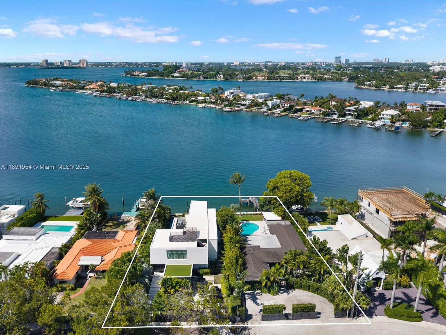 This aerial shot showcases two modern waterfront homes with distinct architectural styles. The property features lush landscaping, a pool, and direct access to the bay, offering stunning views of the surrounding islands and cityscape. The image highlights the property's prime location and luxurious outdoor amenities.