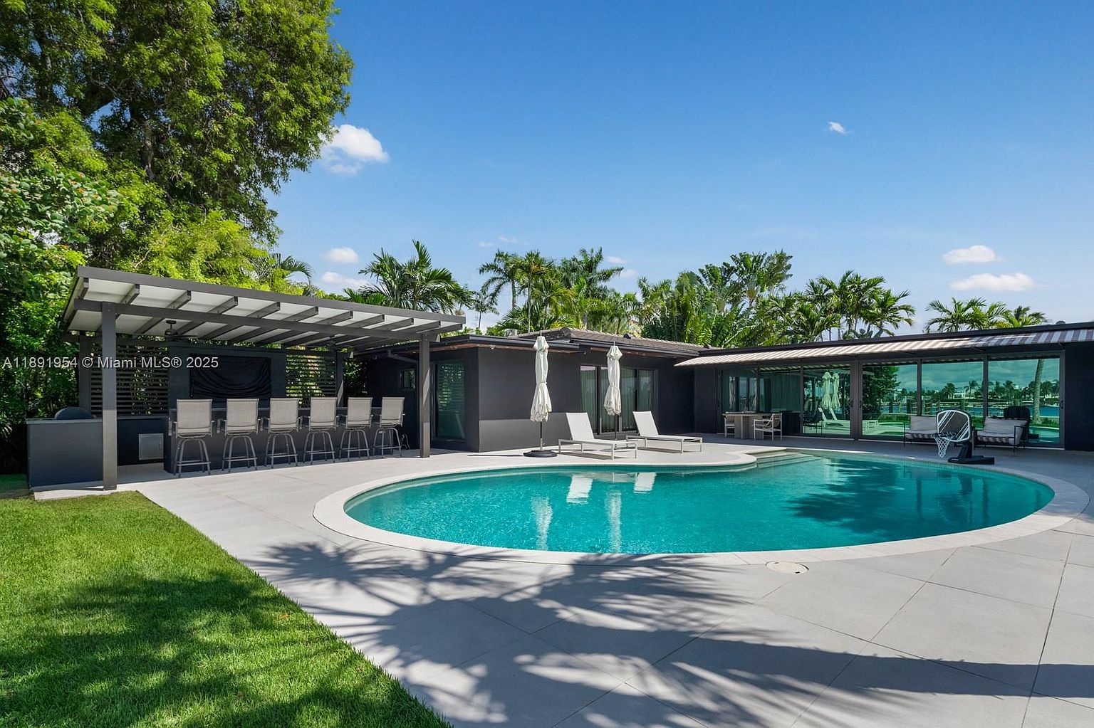 This exterior shot showcases a luxurious backyard pool area. The kidney-shaped pool is surrounded by a concrete patio, with lounge chairs and umbrellas providing relaxation spots. A covered outdoor bar area with seating and a modern home with large windows complete the scene, all set against a backdrop of lush tropical landscaping and a clear blue sky.