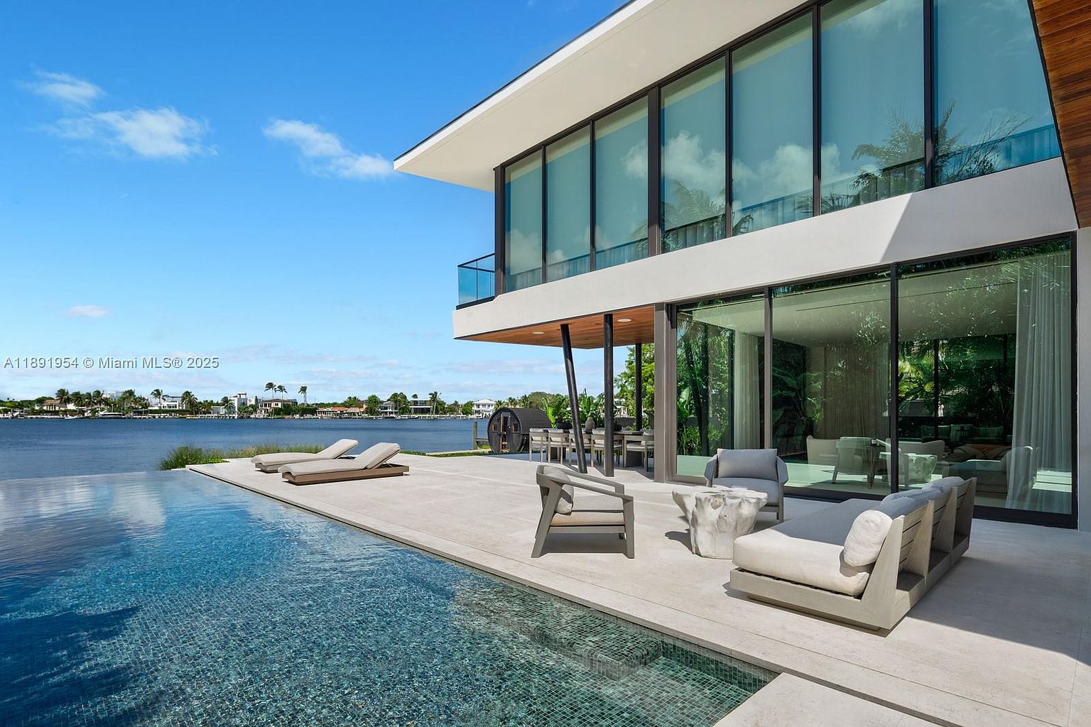 This image showcases a luxurious modern home with a stunning infinity pool that blends seamlessly with the waterfront view. The outdoor living space features contemporary lounge furniture, creating an inviting area for relaxation and entertainment. The architecture emphasizes clean lines and expansive glass windows, offering panoramic views and a sense of openness.