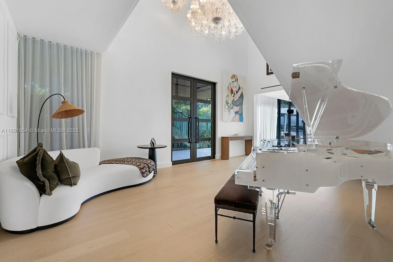 This is a bright and airy living room featuring a modern aesthetic. A clear grand piano sits prominently in the room, complemented by a curved white sofa with decorative pillows and a unique floor lamp. Large windows and a high ceiling with a chandelier contribute to the spacious and luxurious feel of the room.