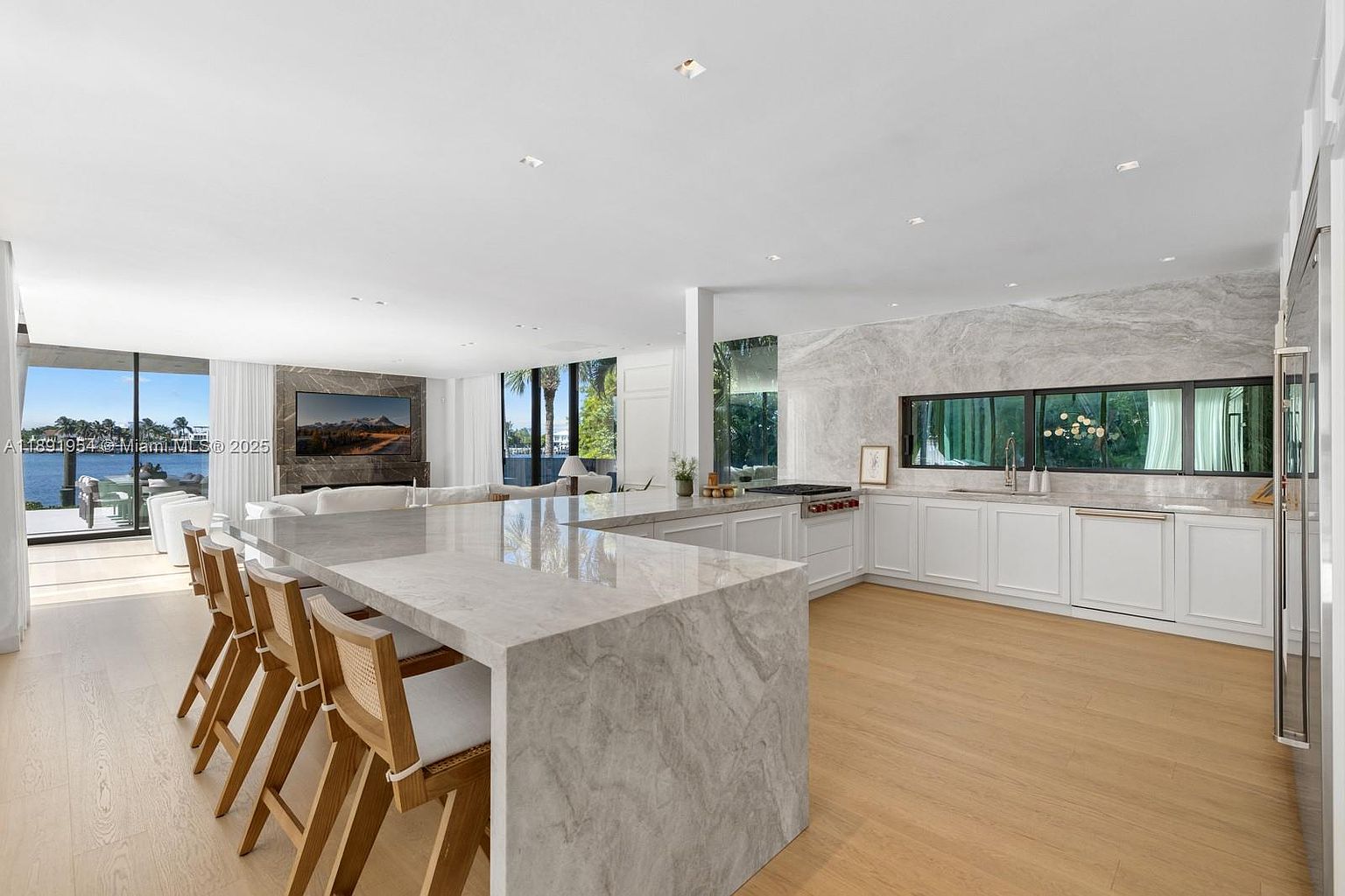 This is a bright, modern kitchen featuring a large marble island with seating, white cabinetry, and marble backsplash. The kitchen opens to a living area with large windows offering water views. The overall impression is luxurious and spacious.