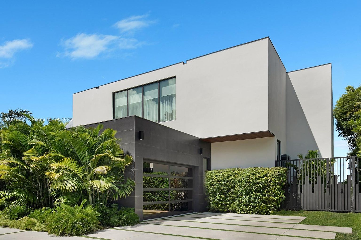 This is a front exterior view of a modern, two-story home. The house features a minimalist design with a white facade, a dark gray garage section with a glass garage door, and lush tropical landscaping. The driveway is made of concrete slabs with grass growing in between, adding a touch of greenery.