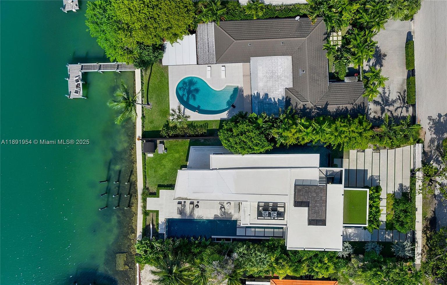 This aerial shot showcases a luxurious waterfront property featuring two distinct homes, each with its own pool and meticulously landscaped grounds. The property boasts a private dock extending into the water, offering direct access for boating and water activities. The architectural style is modern, with clean lines and expansive outdoor living spaces, creating an impression of high-end coastal living.