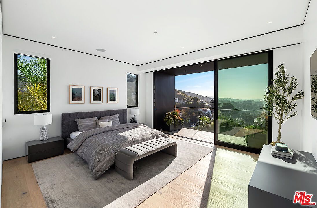 This modern primary bedroom features a neutral color palette with a dark upholstered bed frame and a matching bench at the foot. Large sliding glass doors open onto a private balcony, offering expansive views of the surrounding hills and neighborhood. The room is accented by light wood flooring, a soft area rug, and minimalist decor, creating a serene and sophisticated atmosphere.