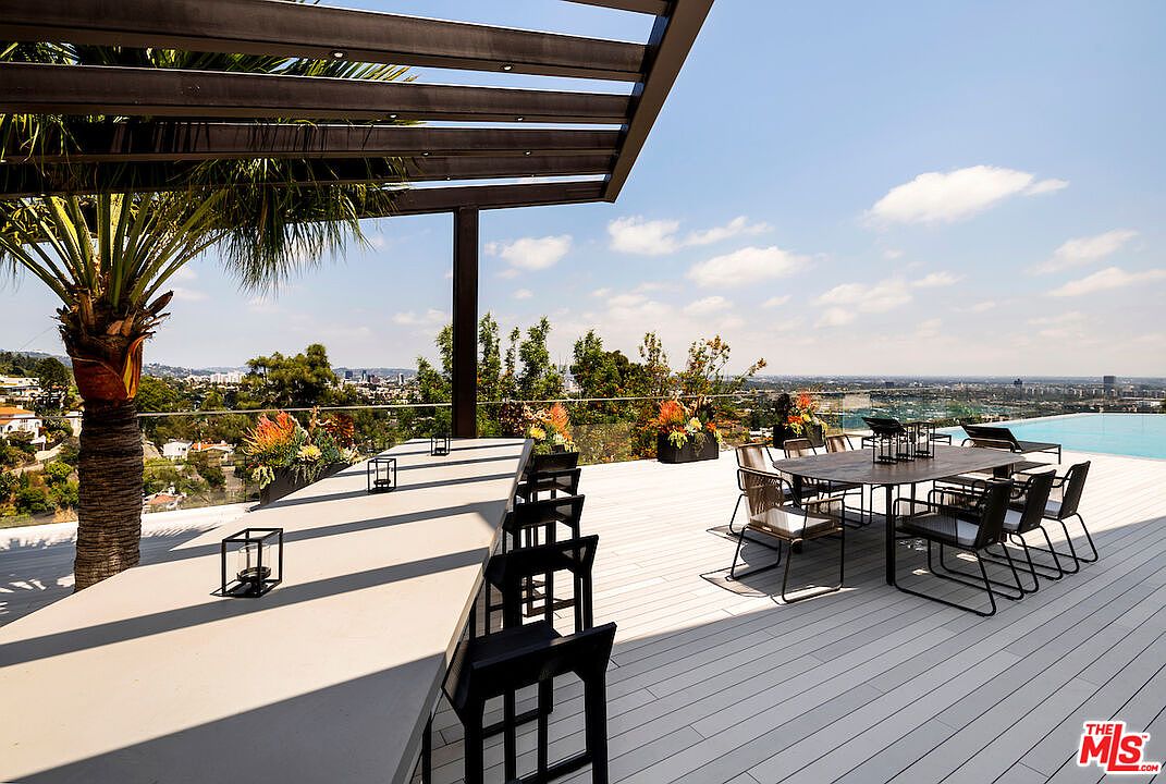 This expansive outdoor deck features a modern pergola structure providing shade over a long, sleek dining table and a separate dining set. The space is elevated, offering panoramic views of the city skyline and surrounding hills, complemented by a glass railing and a nearby infinity-edge pool. A large palm tree adds a touch of tropical elegance to the sophisticated, open-air entertainment area.