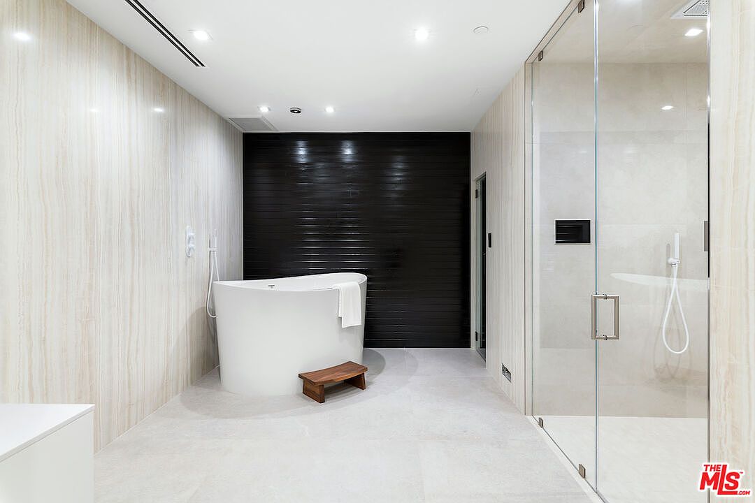 This modern primary bathroom features a minimalist aesthetic with light-toned, vertically-veined wall tiles and a contrasting dark, horizontal-slatted accent wall behind a freestanding soaking tub. A glass-enclosed walk-in shower sits to the right, while a small wooden stool provides a functional accent in front of the tub. The perspective is centered, highlighting the clean lines and high-end, spa-like atmosphere of the space.
