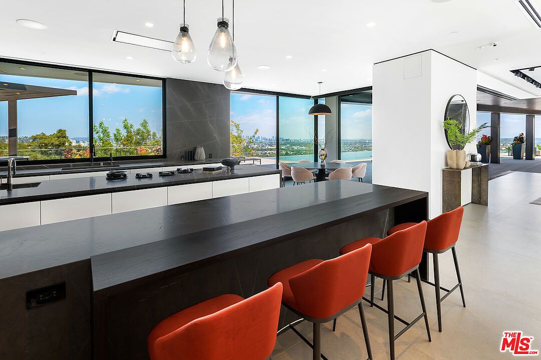 This sleek, modern kitchen features a large, dark-toned island with integrated seating for four, complemented by vibrant orange bar stools. The space is defined by minimalist white cabinetry, dark stone countertops, and expansive glass walls that provide panoramic views of the city skyline. Pendant lights hang above the island, while a separate dining area is visible in the background, creating an open-concept layout perfect for entertaining.
