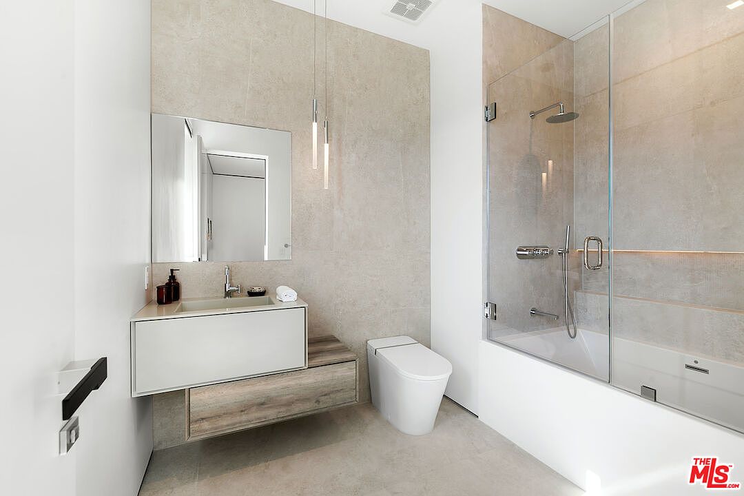 This modern bathroom features a minimalist aesthetic with neutral stone-look wall tiles and a floating vanity. The space includes a sleek, wall-mounted toilet and a glass-enclosed shower area with a rainfall showerhead. Two slender pendant lights hang above the vanity, adding a contemporary touch to the clean, bright design.
